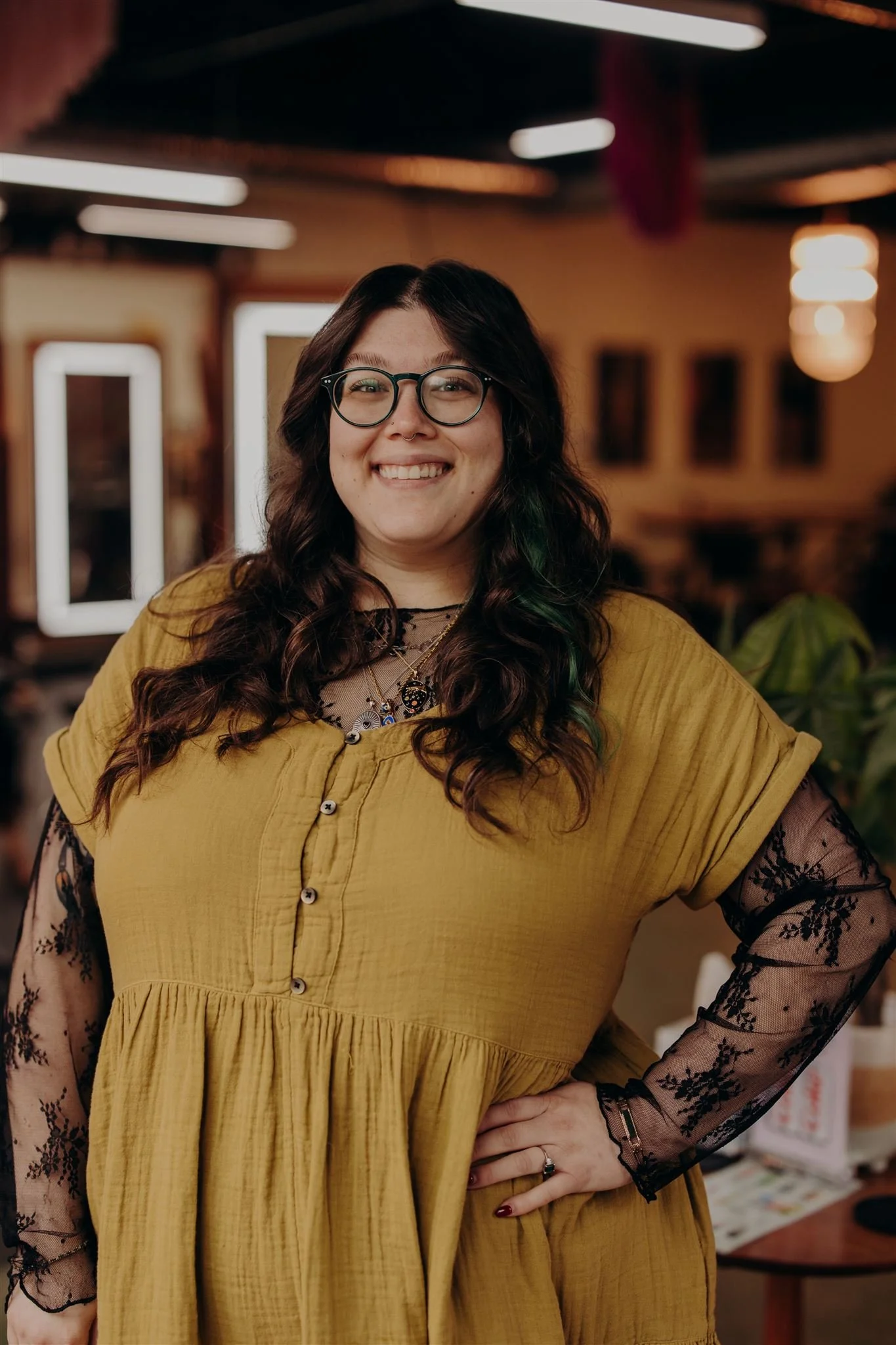 A woman with dark curly hair, glasses, and a septum piercing smiling against a yellow background. She is wearing a black sleeveless top, layered necklaces, and a checkered skirt with a black belt.