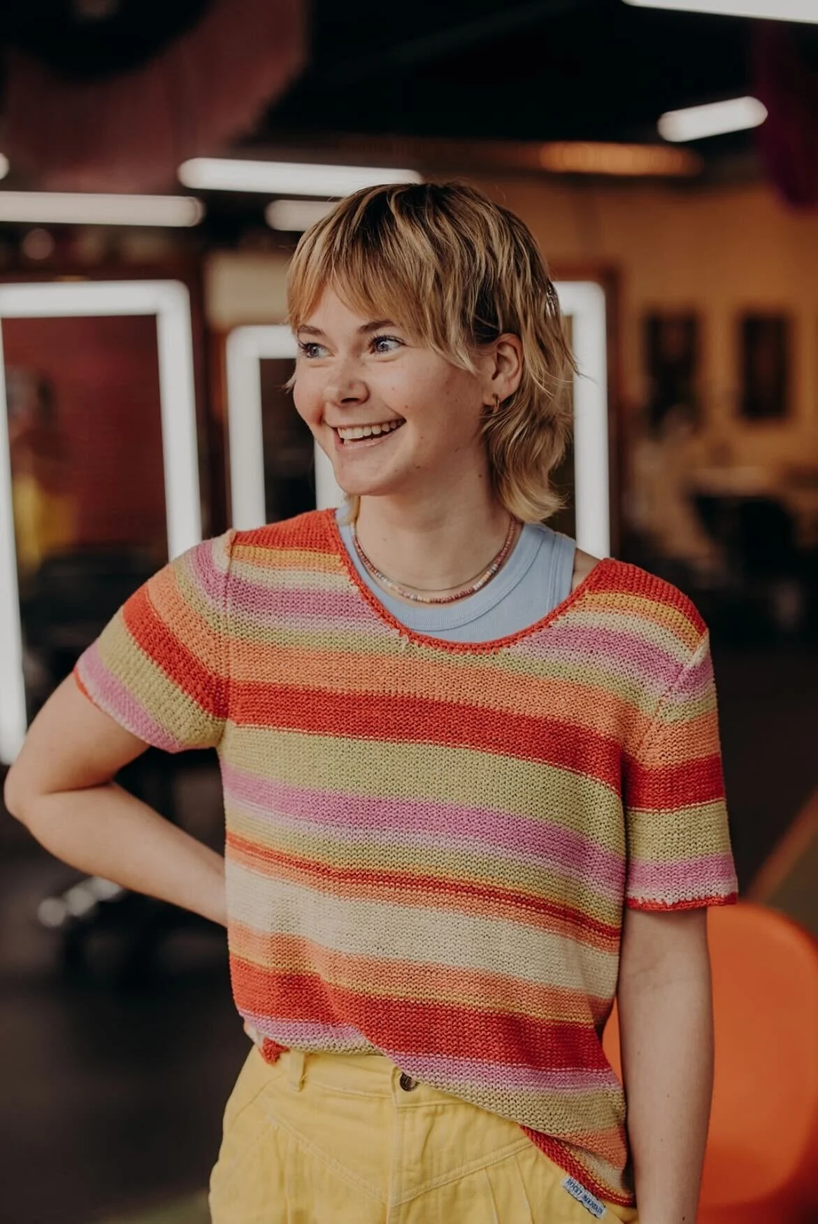 A young woman with blonde hair, wearing a sleeveless embroidered vest over a green crop top, standing in front of a red brick wall and smiling.