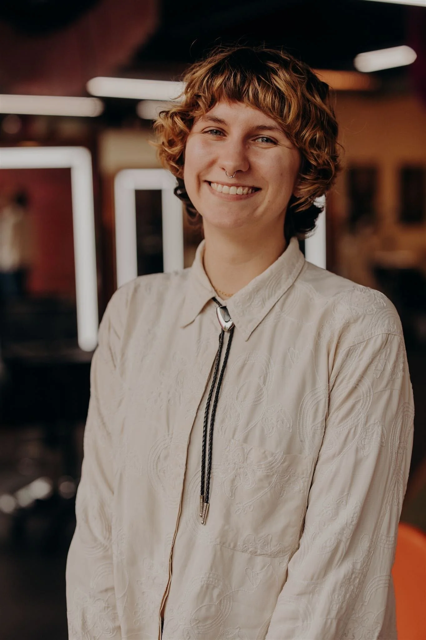 A young person with short, curly dark hair, wearing a green patterned shirt, gold necklaces, and earrings, smiling at the camera against an orange background.