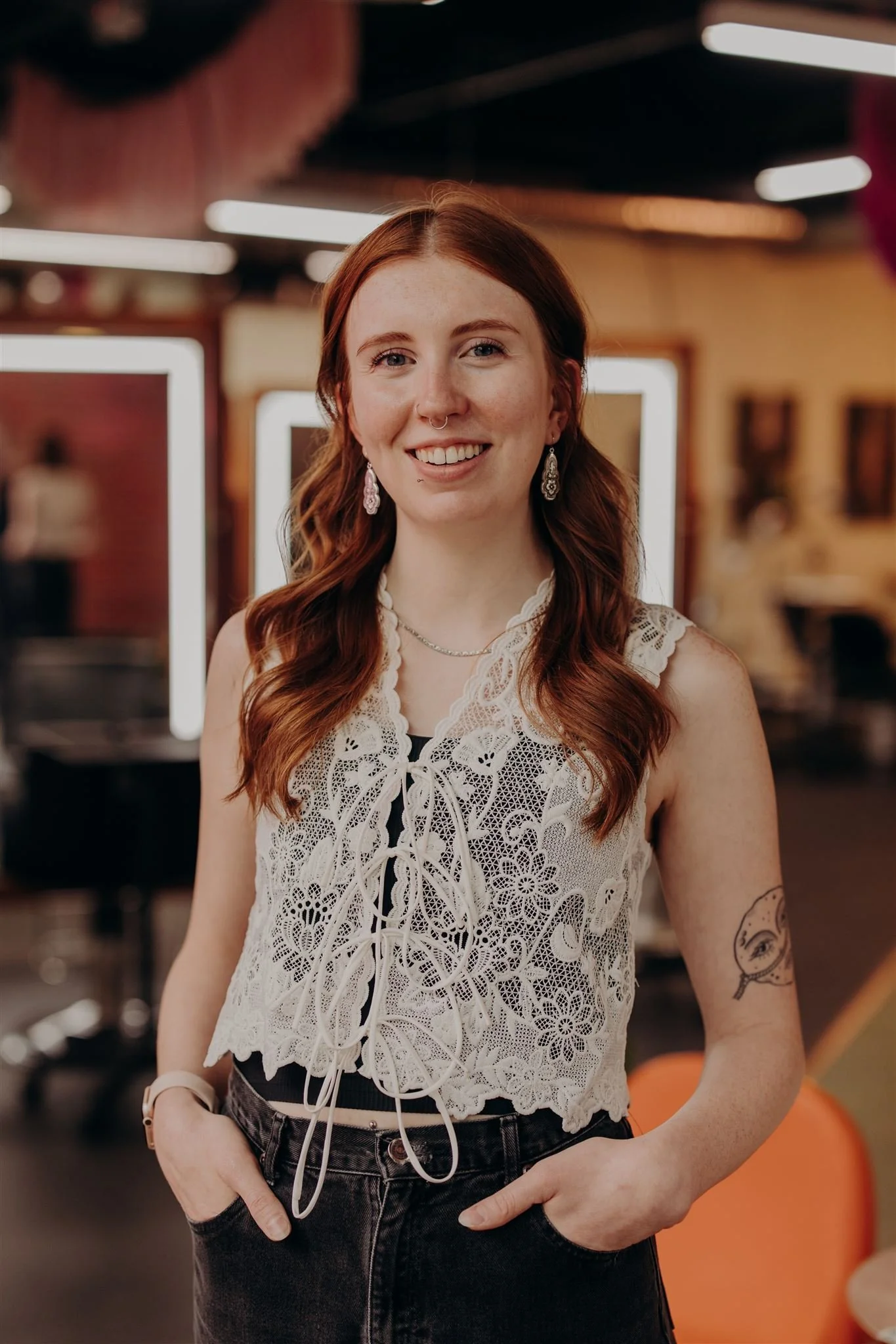 A young woman with long, red hair wearing a black sleeveless top, jewelry including earrings and necklaces, a belt with metal loops, and a smartwatch, standing against a dark background.