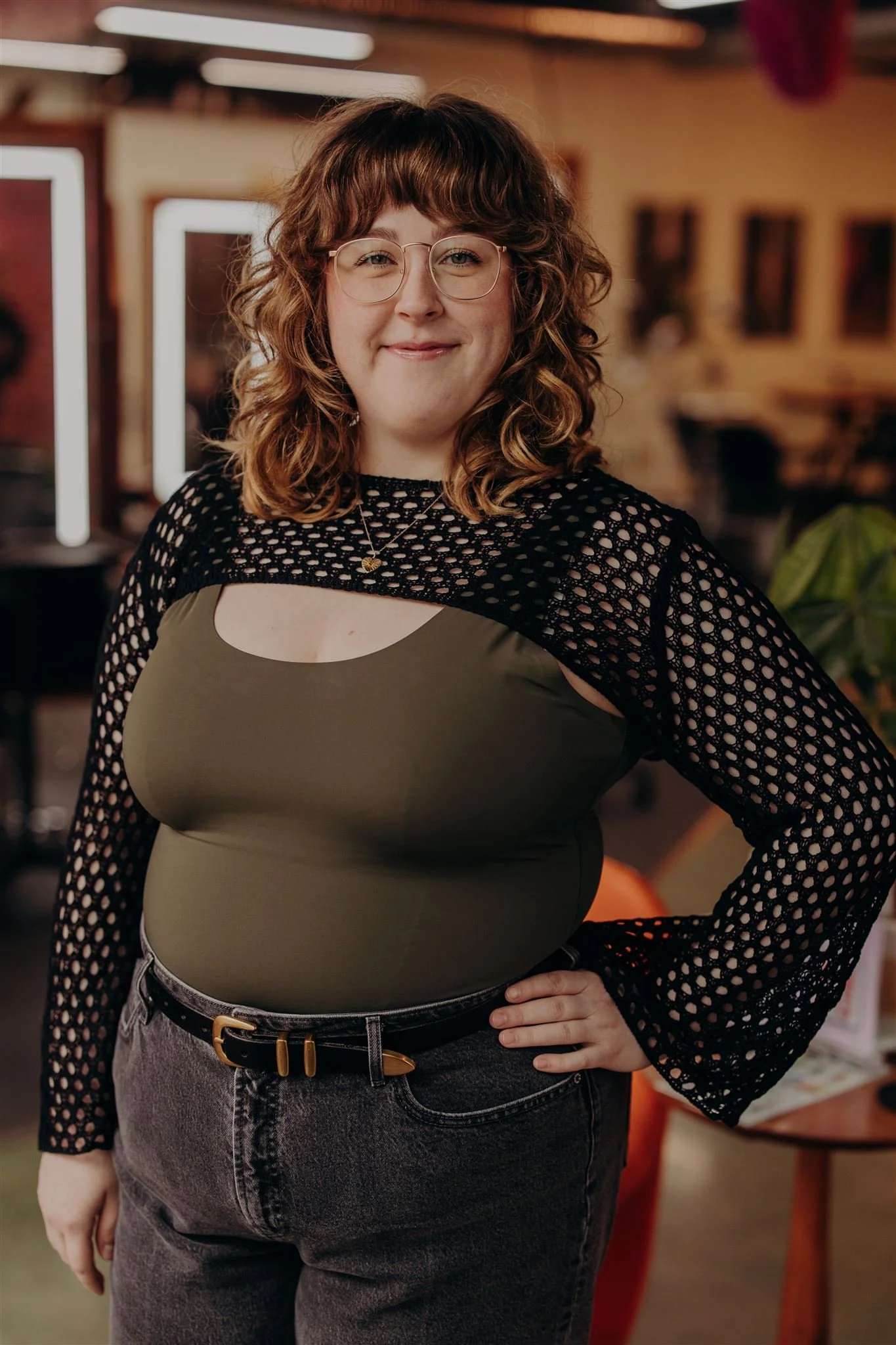 A woman with curly, ombre yellow and orange hair, wearing glasses, a black T-shirt, a denim vest, and a white skirt, standing against a black background.