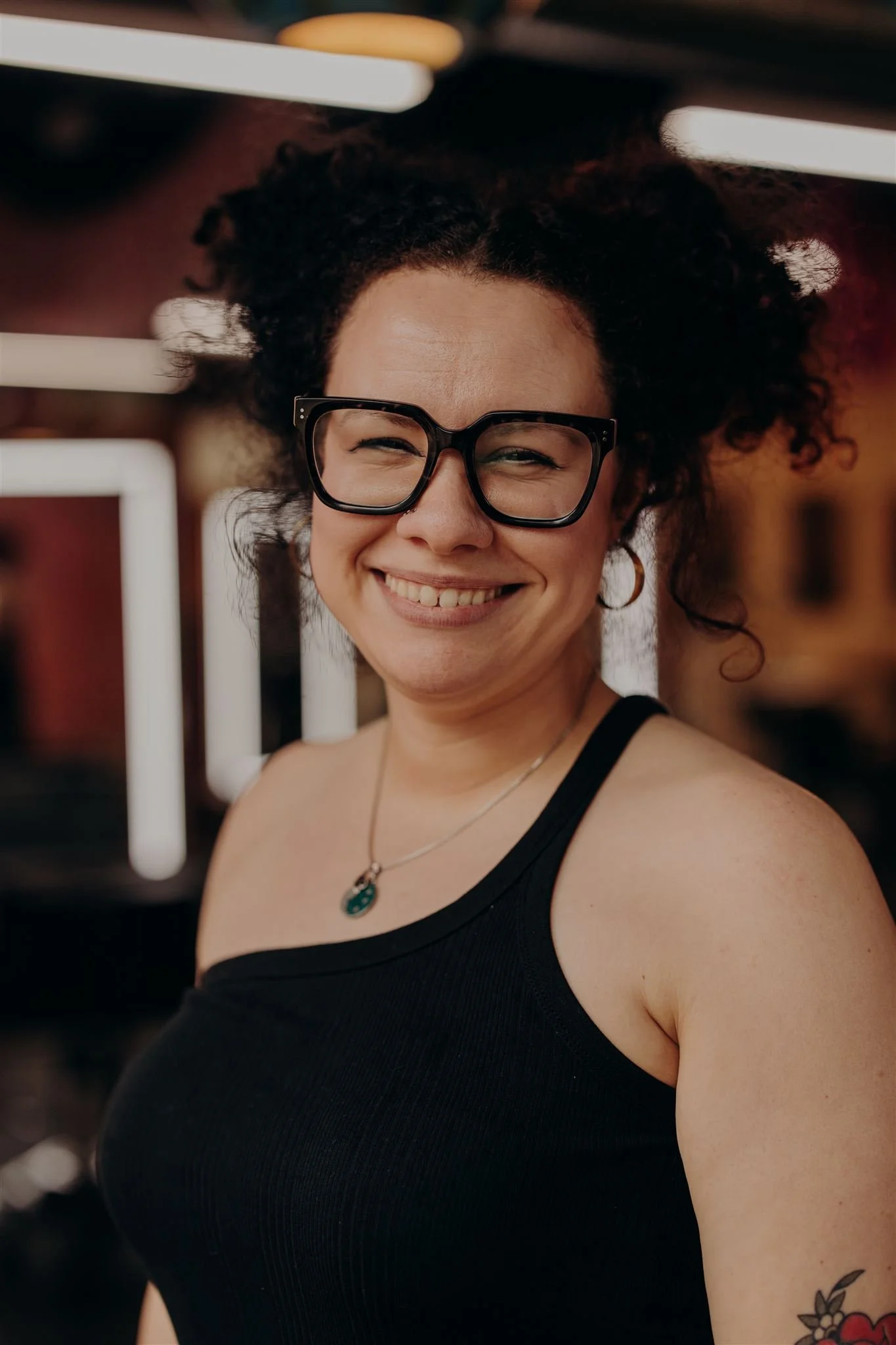A woman with curly hair and glasses smiling, wearing a black blouse, red plaid pants, and black loafers with studs, standing against a plain beige background.