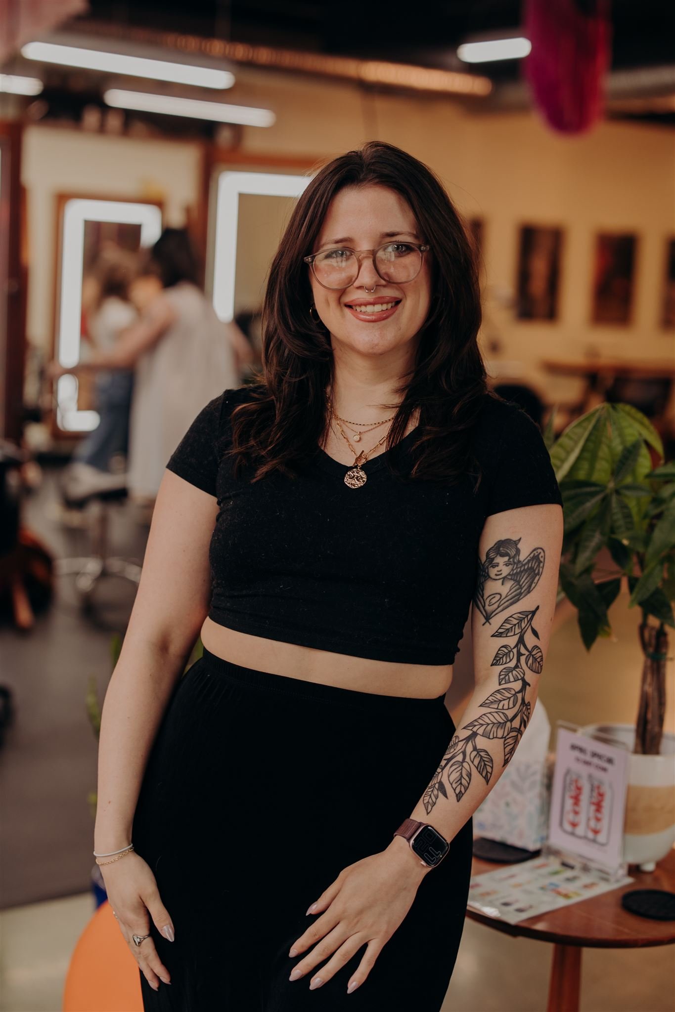 A smiling young woman with short brown hair, wearing glasses, a nose ring, gold earrings, colorful necklaces, a black sleeveless top, and beige pants, standing in front of a red brick wall.