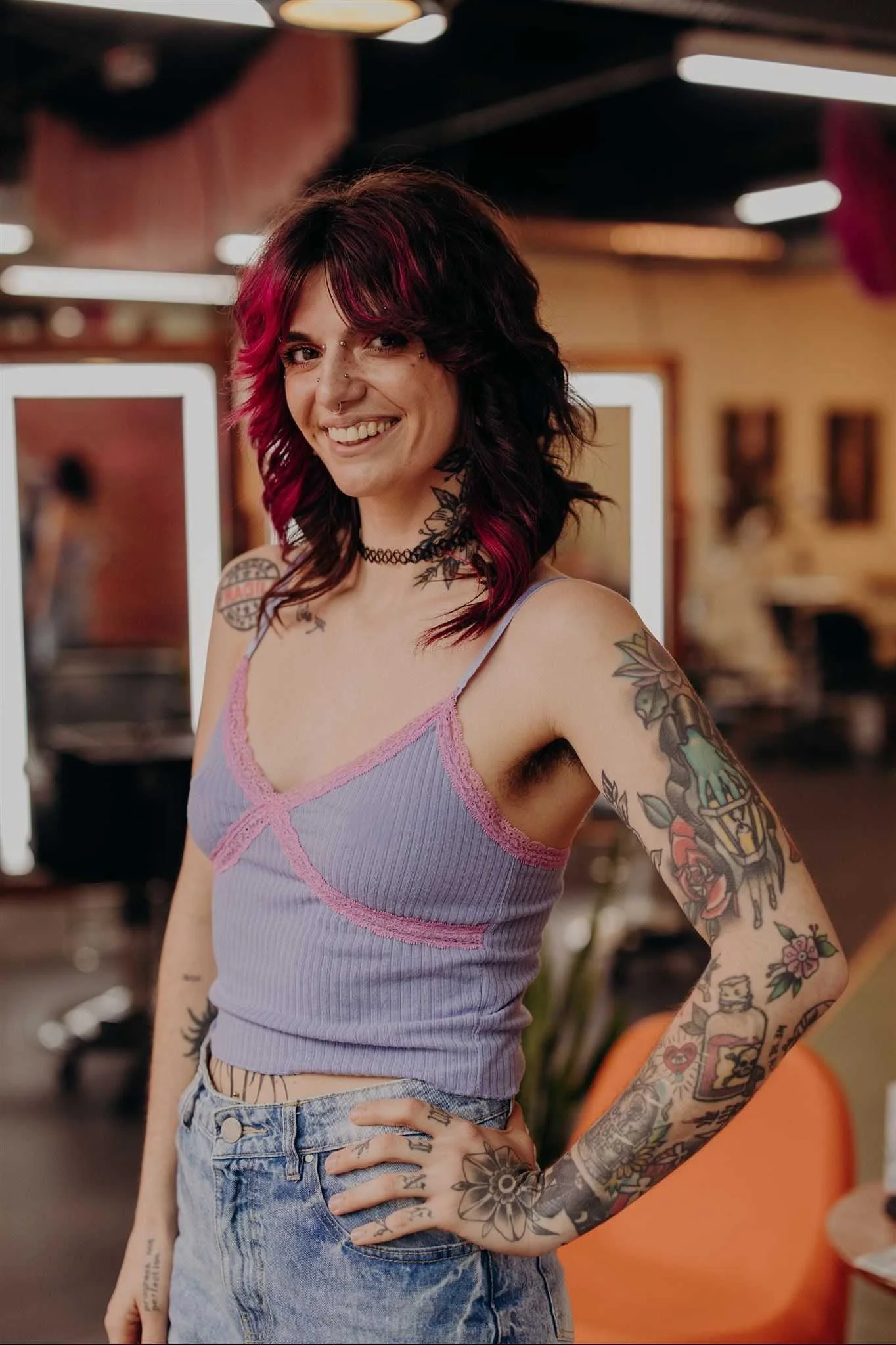 A person with short dark hair with orange highlights, tattoos on their arms and chest, smiling and posing in front of an orange background.