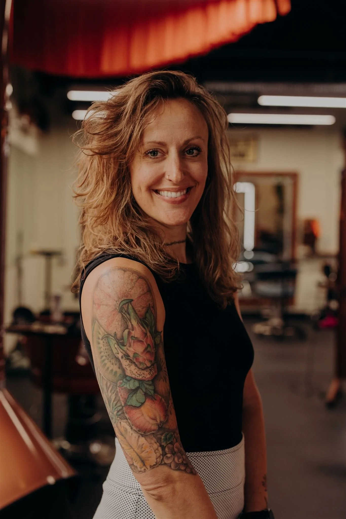 A smiling woman with shoulder-length brown hair wearing a black sleeveless top, layered necklaces, and a nose ring, posing against a blue background.