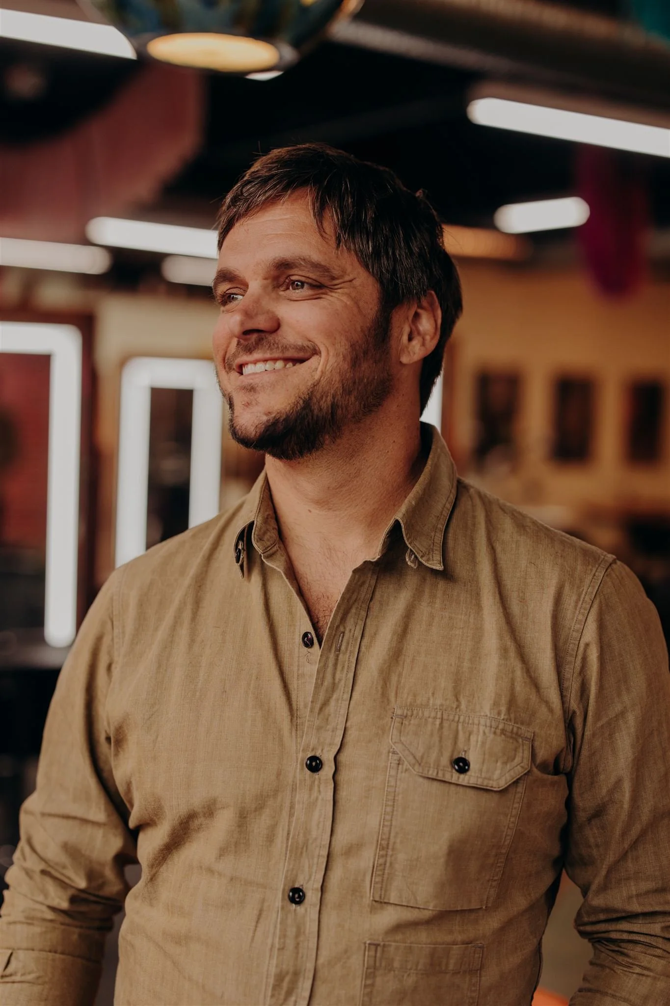 A close-up portrait of a smiling man with short brown hair, light skin, and a beard, wearing a black shirt with intricate patterns, against a dark background.