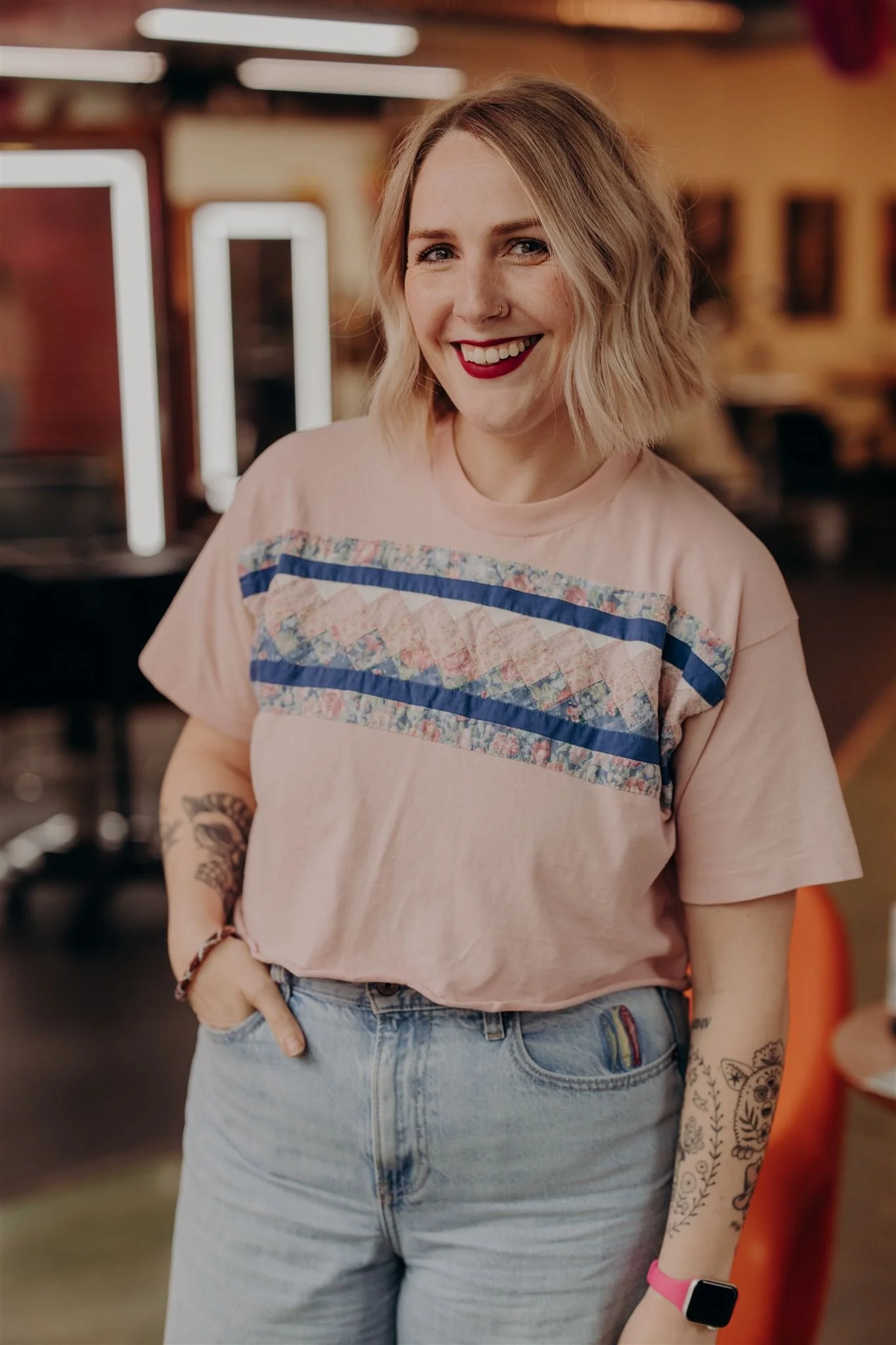 A woman with short blonde hair wearing a blue and white striped shirt, tattoos on her arms, and an Apple Watch, standing against a pink background.