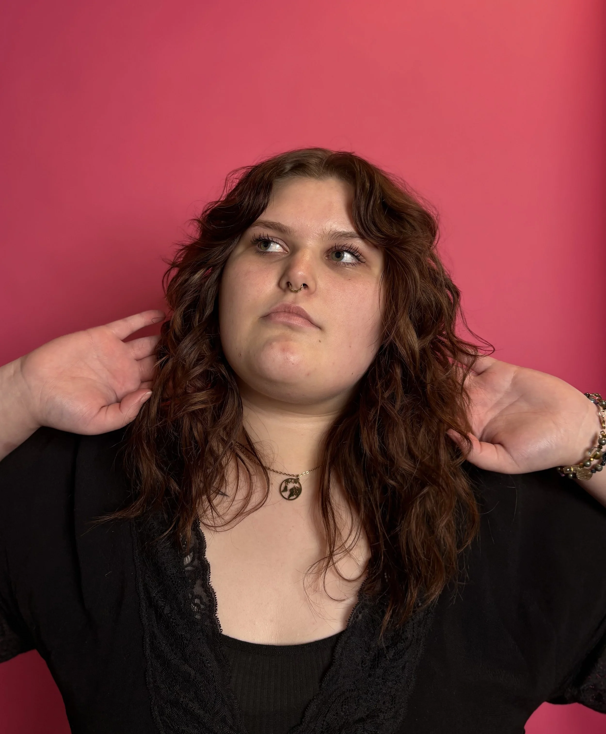 Young woman with wavy brown hair, wearing a black top, jewelry, and a septum piercing, posing against a pink background.