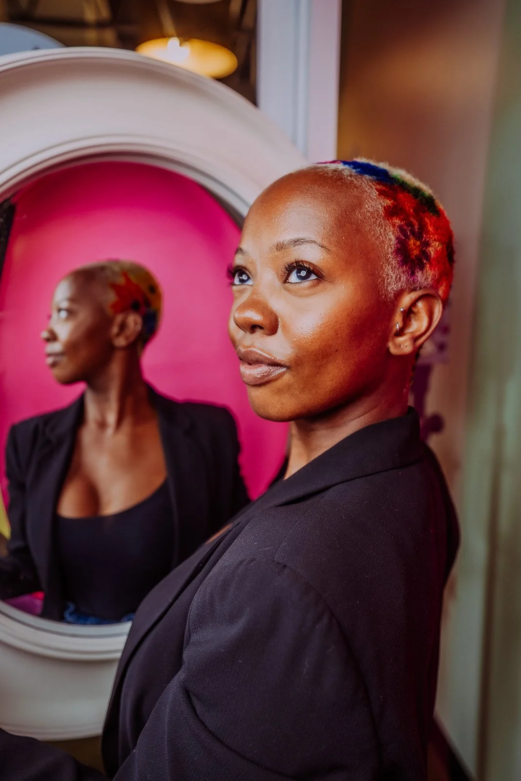 Portrait of a woman with colorful short hair looking away, with her reflection in a mirror showing her in profile against a pink background.