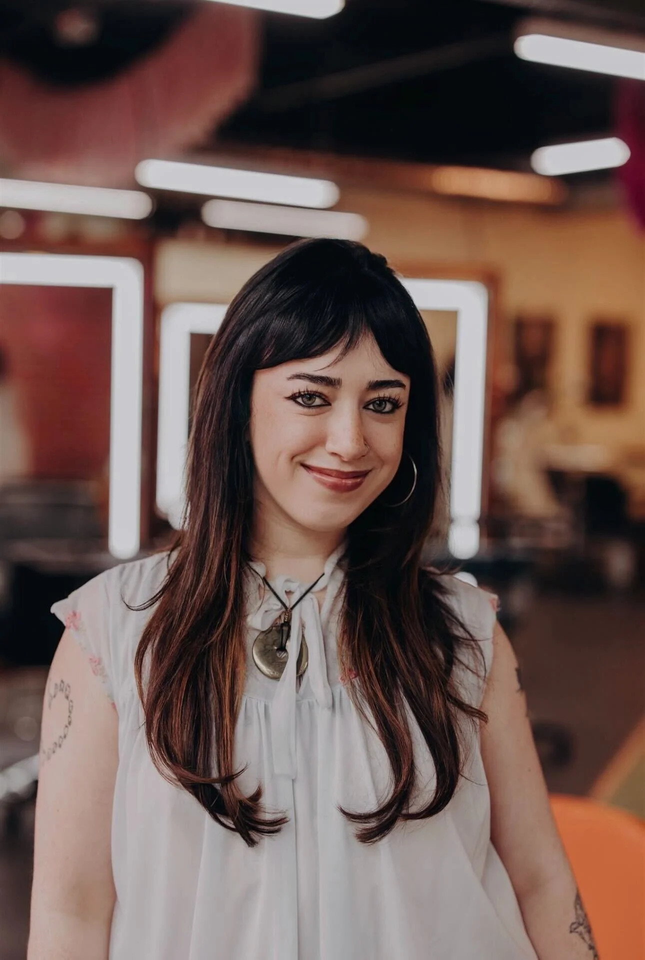 A young woman with long, wavy dark hair and light-colored eyes, wearing a black lace top and a large iridescent gemstone necklace, standing in front of a red brick wall.