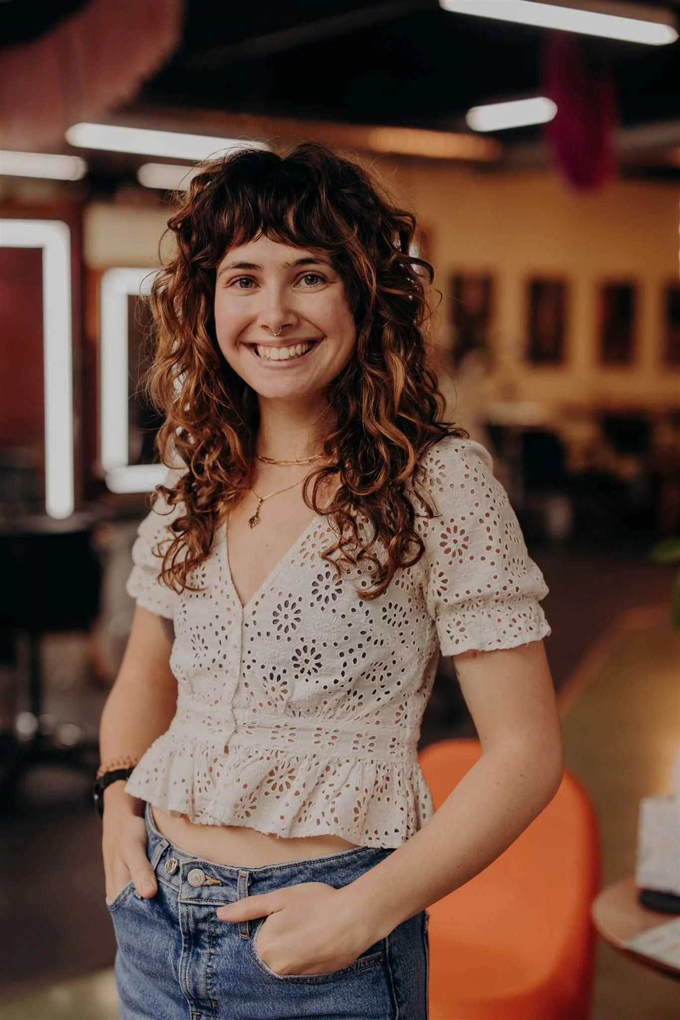 A young woman with curly brown hair, smiling and standing against a red brick wall, wearing a light pink ribbed crop top, blue jeans, a black watch, and a necklace.