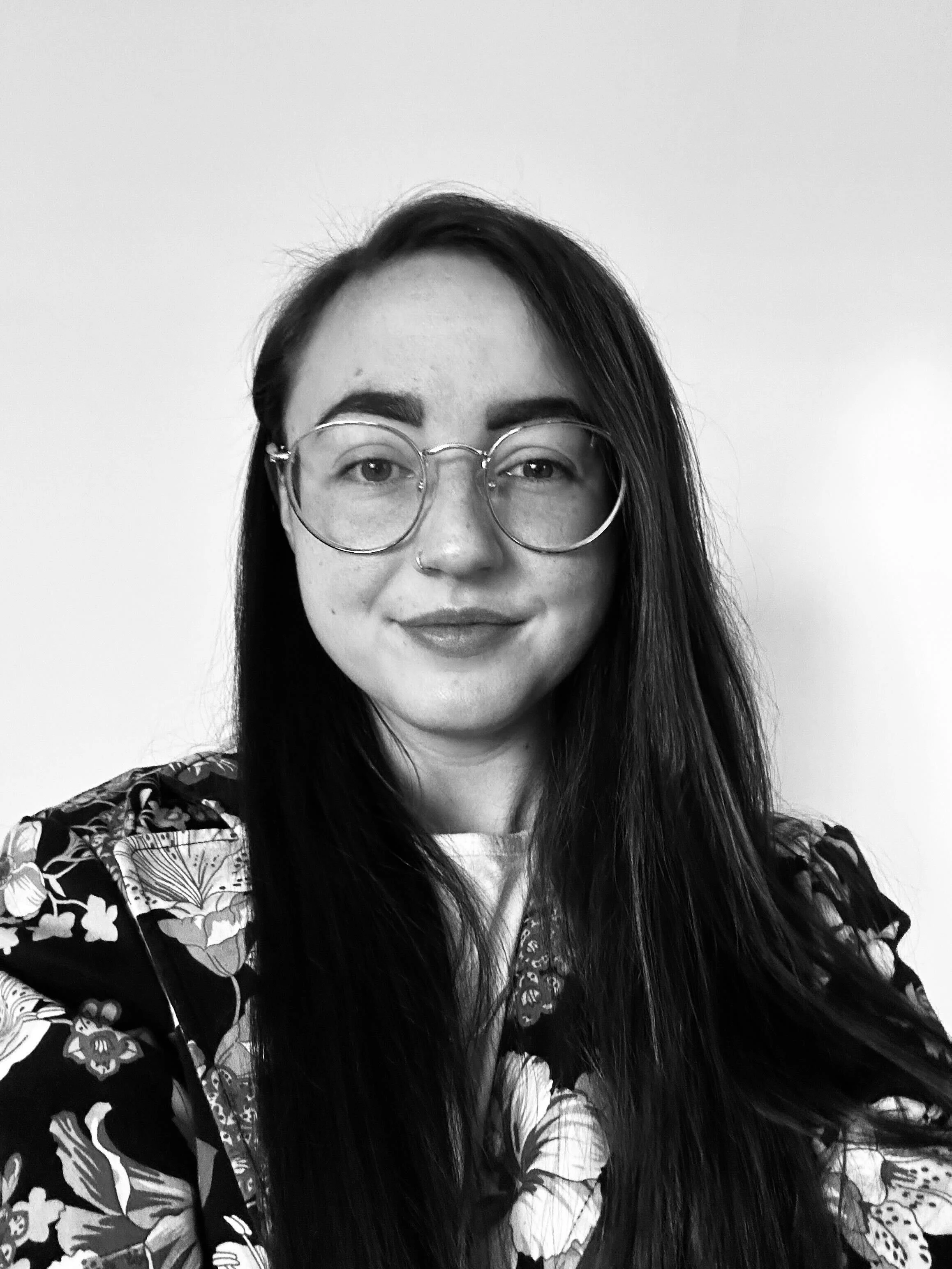 A black and white portrait of a young woman with long dark hair, wearing round glasses and a floral patterned top, smiling slightly in front of a plain white background.