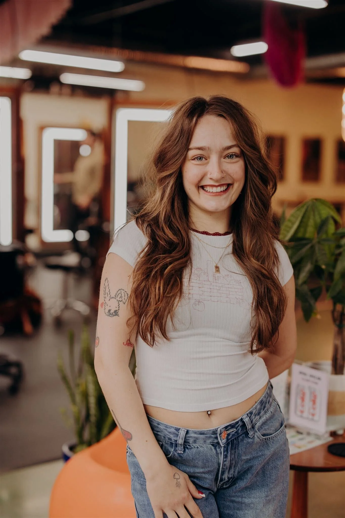 A smiling young woman with long brown hair, wearing a denim top, layered gold necklaces, hoop earrings, and a leopard print belt, standing in front of a colorful retro patterned wall.