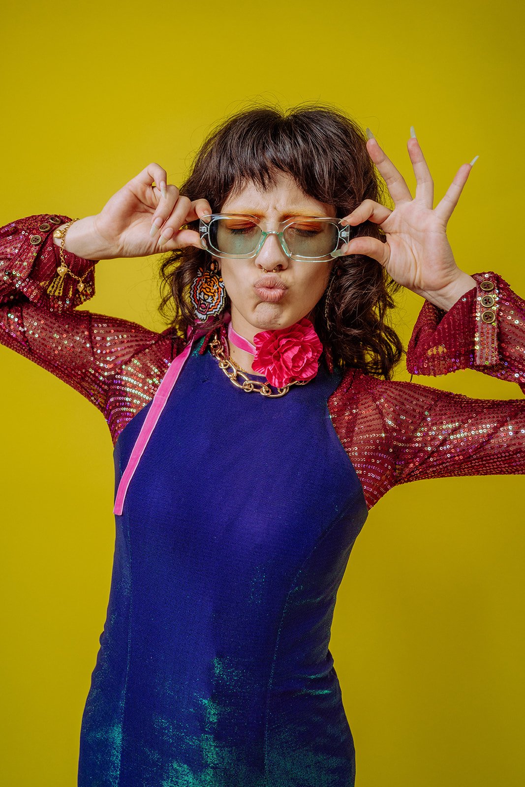 Woman wearing colorful clothing and accessories, making a playful face and gestures against a yellow background.