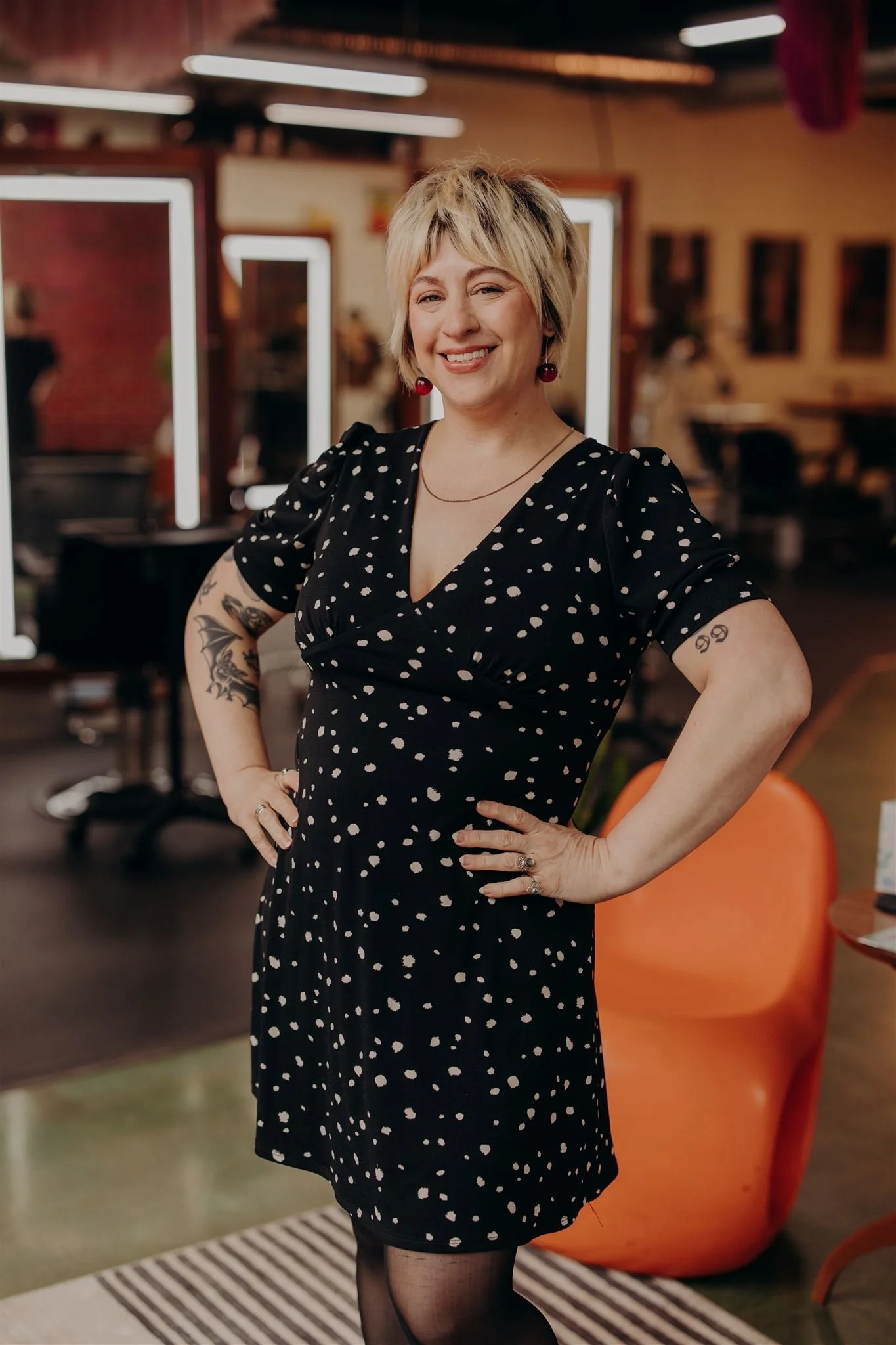 A woman with short pink hair, smiling and wearing a black sleeveless top, standing in front of a wall decorated with yellow bananas.