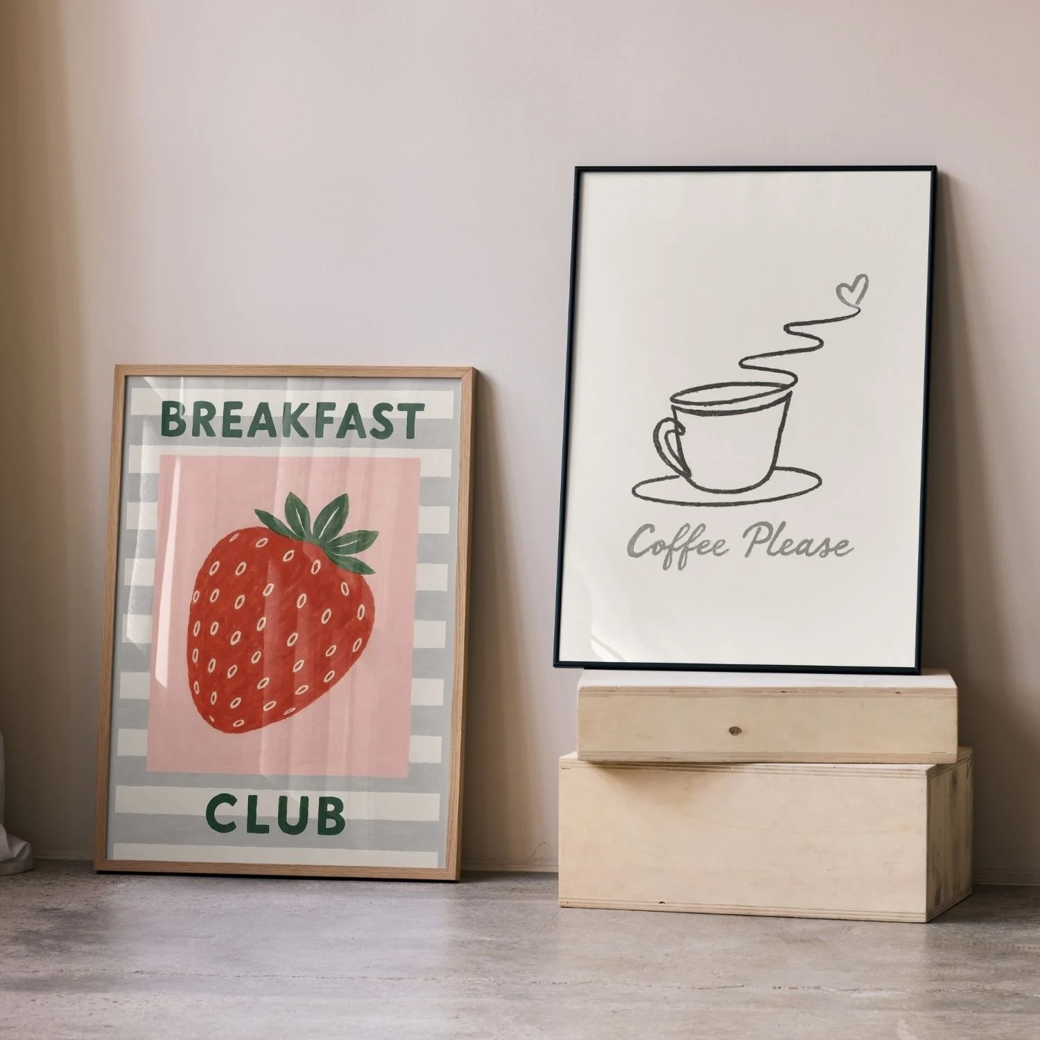 Two framed posters, one with a large strawberry illustration and the words 'Breakfast Club' in green text, the other with a simple line drawing of a coffee cup and the words 'Coffee Please' in gray text, resting on a wooden platform against a white wall.