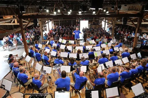 Large orchestra performing in a venue with a conductor leading, and audience watching.