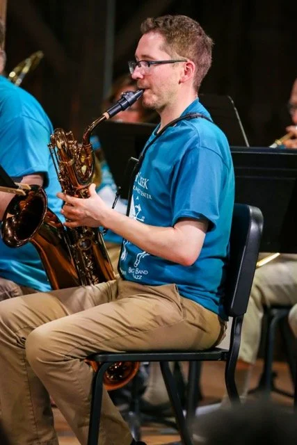 A man wearing glasses and a blue t-shirt playing a black saxophone in an orchestra.