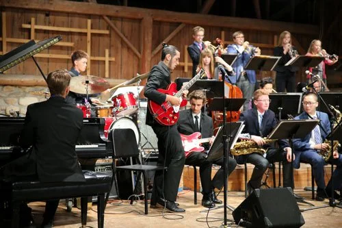 A group of young musicians performing on stage with a piano, drums, guitars, and wind instruments.