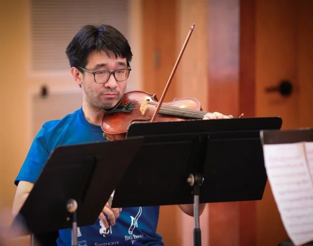 A man with glasses and dark hair is playing a violin while reading sheet music, in a room with wooden walls.