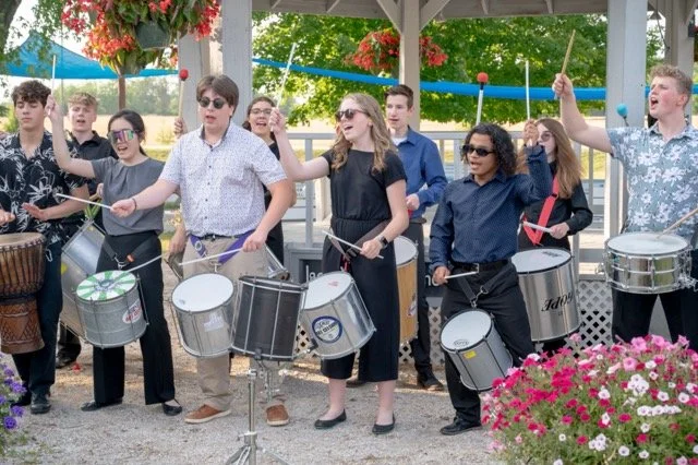 Group of young people playing drums and percussion instruments outdoors with colorful flowers.