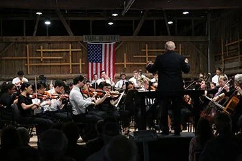 An orchestra performing on stage with a conductor in front, American flag in the background, and wooden walls.