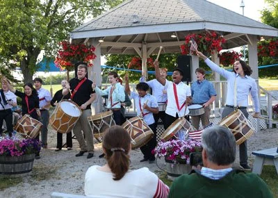 A group of musicians playing drums and percussion instruments in a park pavilion during an outdoor event, with an audience watching.