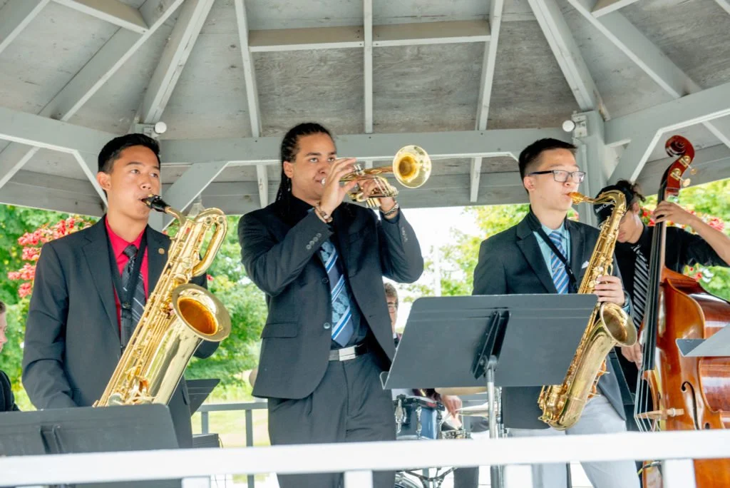 Four young musicians in suits playing jazz instruments in an outdoor pavilion, with blooming trees in the background.