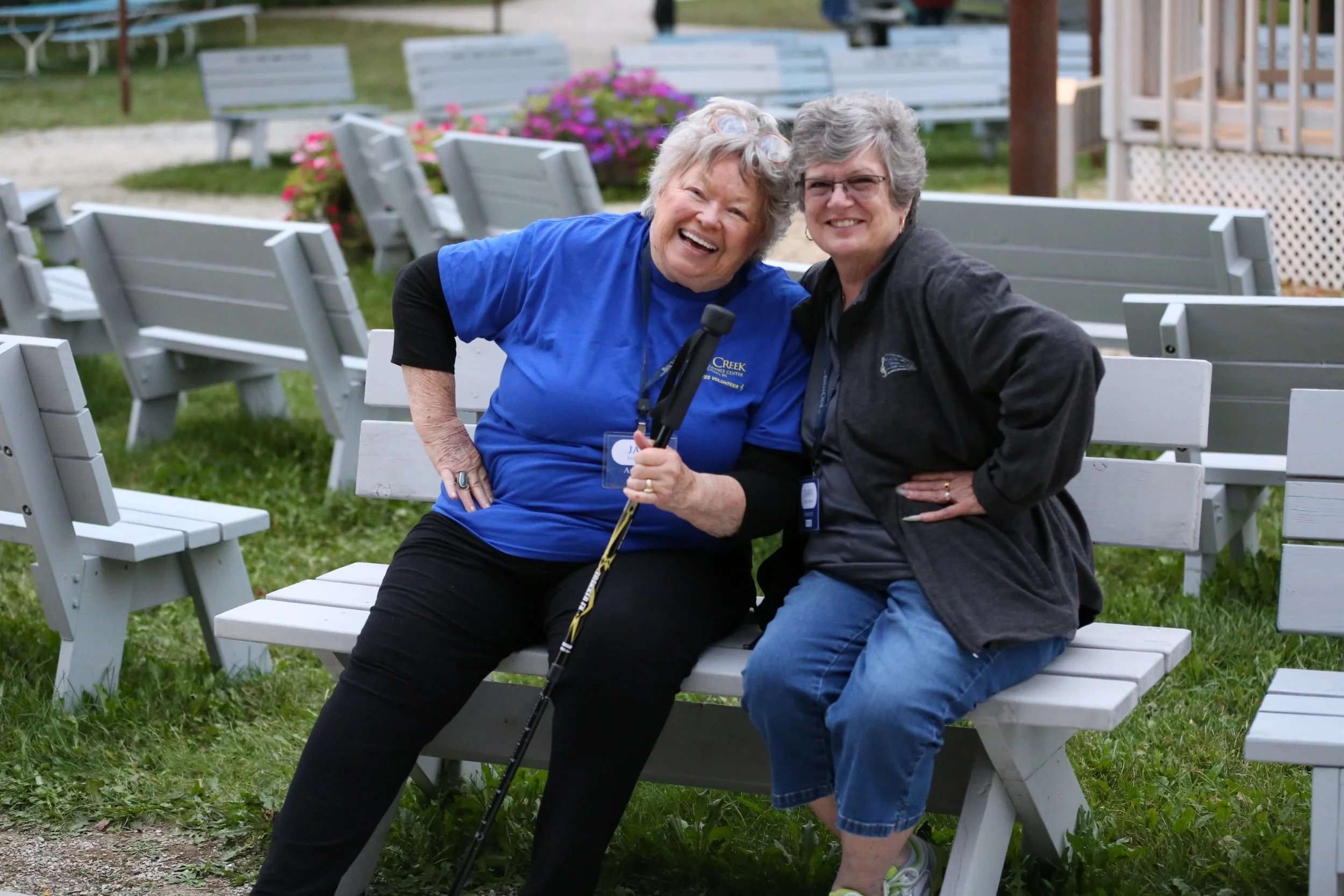 Two women smiling and sitting on a park bench outdoors. One woman is holding a cane and wearing a blue shirt, while the other is wearing a black jacket and blue jeans. They appear to be enjoying a cheerful moment together.