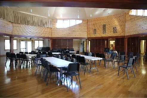 Empty conference or banquet hall with white tables and black chairs, wood-paneled walls, large windows, and hardwood floors.