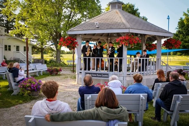 A band performing on an outdoor gazebo stage with audience seated on benches, surrounded by trees and flowers.