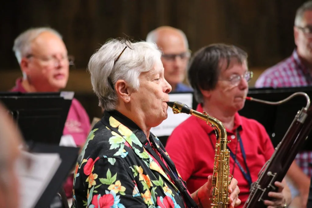 An elderly woman with short gray hair and glasses, wearing a floral shirt, playing a gold-colored saxophone during a group rehearsal, with other musicians and music stands in the background.