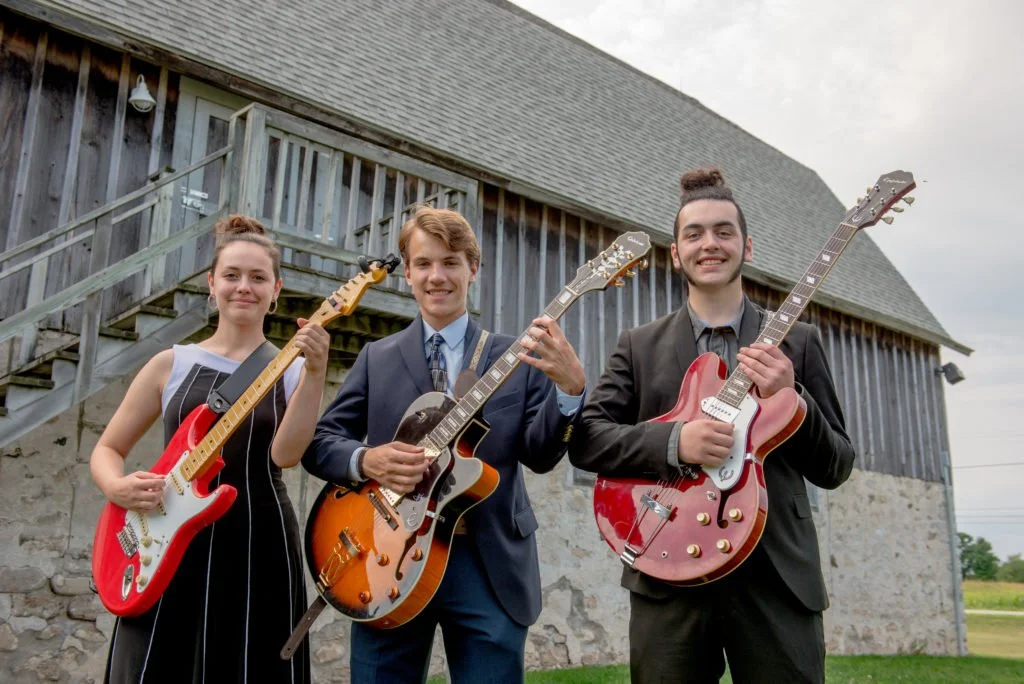 Three young musicians in formal attire holding electric guitars outside a rustic building with a wooden barn and stone foundation.