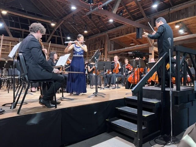 Orchestra performing on stage in a rustic wooden barn, with musicians playing string and wind instruments.