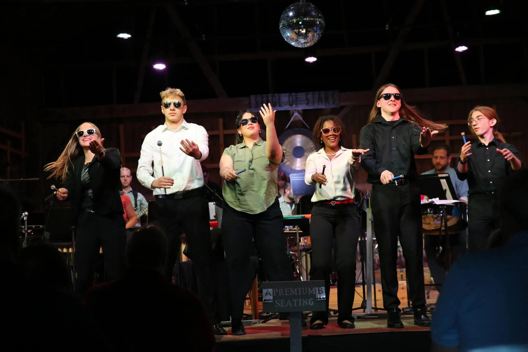A group of six people on stage wearing sunglasses, singing or performing with microphones in a music or theater setting, with a band or musicians in the background and a disco ball overhead.