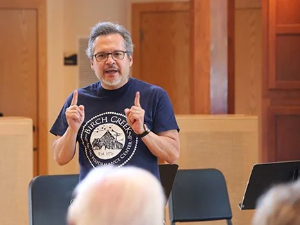 Man with glasses and gray hair, wearing a navy T-shirt, speaking and gesturing with both index fingers in front of an audience in a conference room.