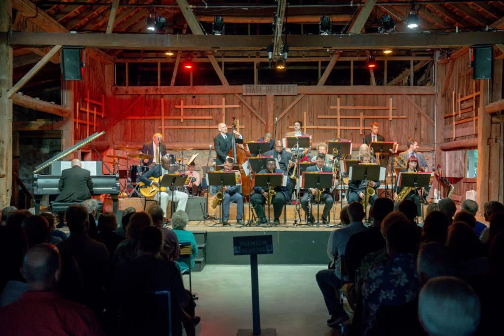 Jazz band performing on stage inside a wooden barn, with audience seated in front. The band includes a pianist, guitarist, bassist, drums, and brass players. Stage lights and a banner reading 'Stage of Stars' are visible.