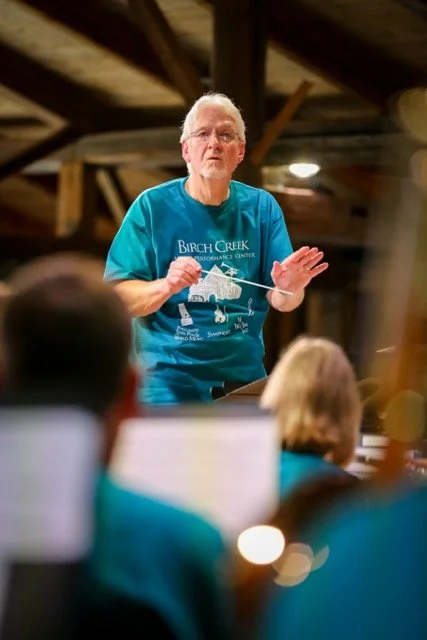 An older man with glasses and white hair conducting or speaking to a group of people in a rustic wooden setting. He is wearing a blue T-shirt with 'Burch Creek' printed on it.