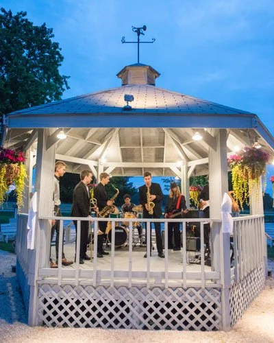 A band of musicians performing inside a white gazebo decorated with hanging flowers and lights