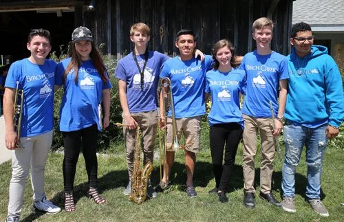 Seven teenagers standing outdoors on grass, wearing matching blue shirts with 'Beach Creek' logo, some with musical instruments, smiling at the camera.