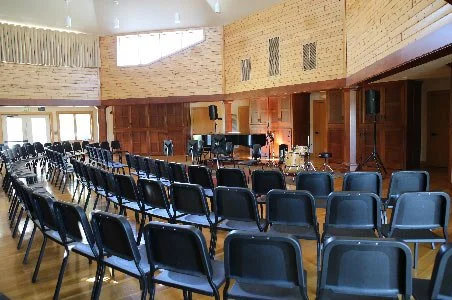 Empty chairs arranged in rows in an auditorium with a stage and piano, wood-paneled walls, and large windows.