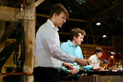Two young men playing xylophones in a rustic venue with wooden beams and stage lighting.