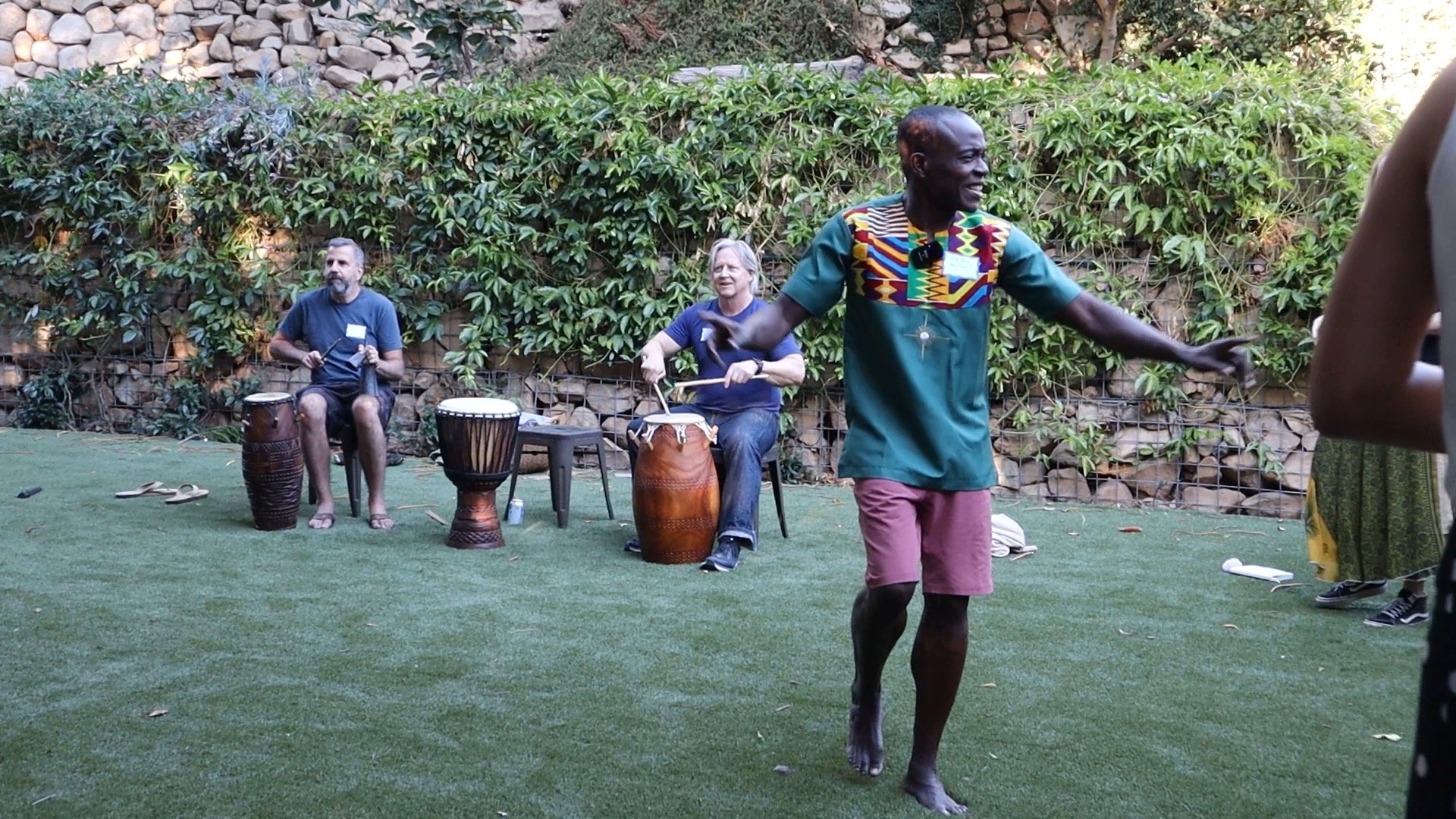 A group of people, one man dancing and two men playing drums, in a backyard with a green grassy area, surrounded by a stone and greenery wall.