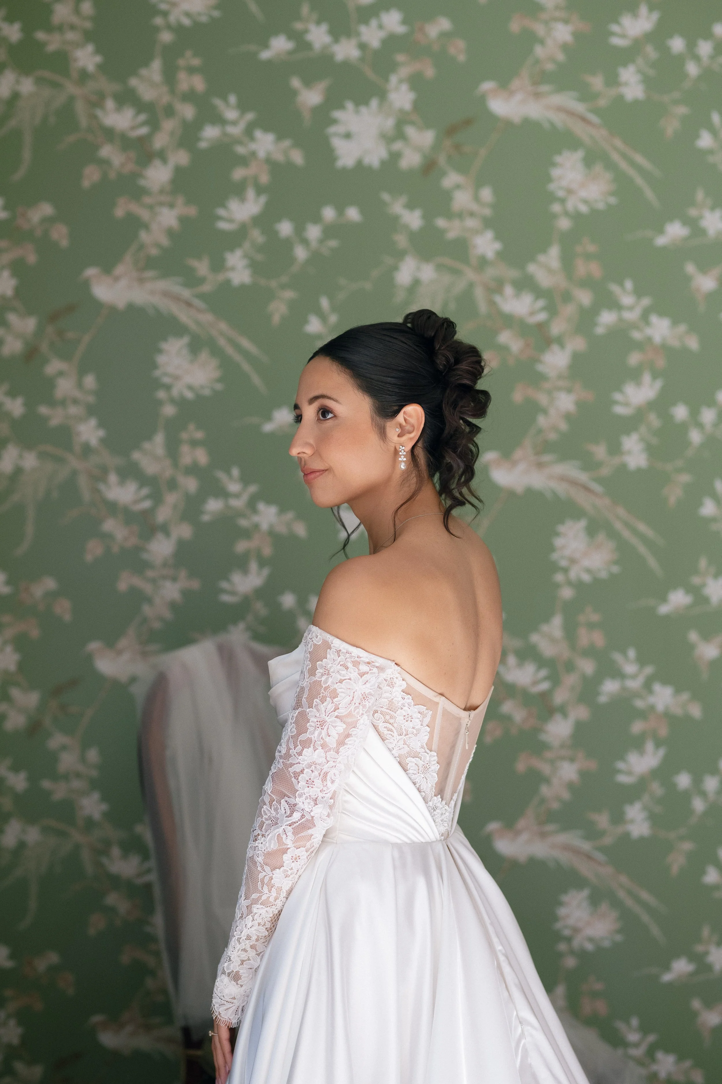 A woman in an off-the-shoulder white lace wedding dress standing sideways against a green floral wallpaper background.