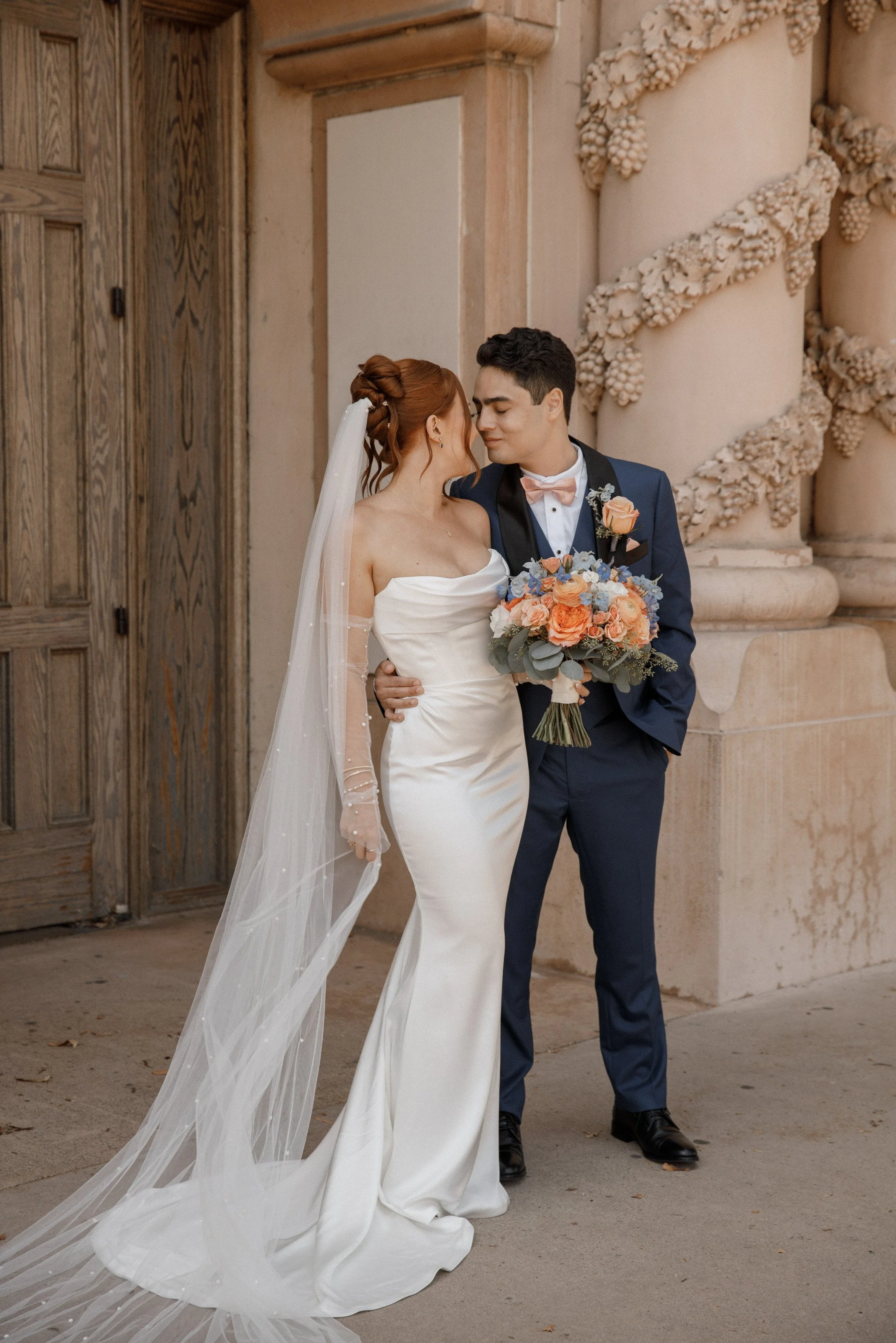 Bride and groom on their wedding day standing close together outdoors, with the bride wearing a strapless white gown with a long veil, and the groom in a navy blue suit with a pink bow tie, holding a bouquet of pink, peach, and purple flowers, standi