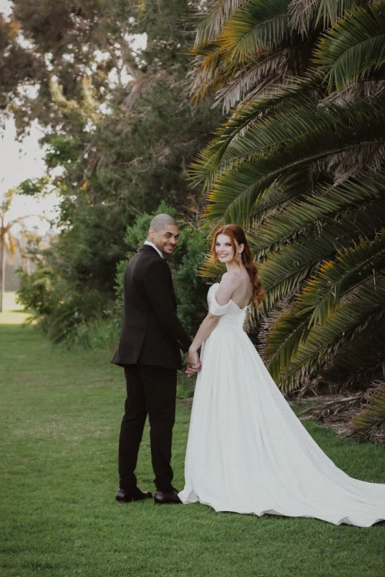 A bride and groom holding hands outdoors near large palm leaves, smiling and looking back at the camera, wedding attire, grassy area, daylight.