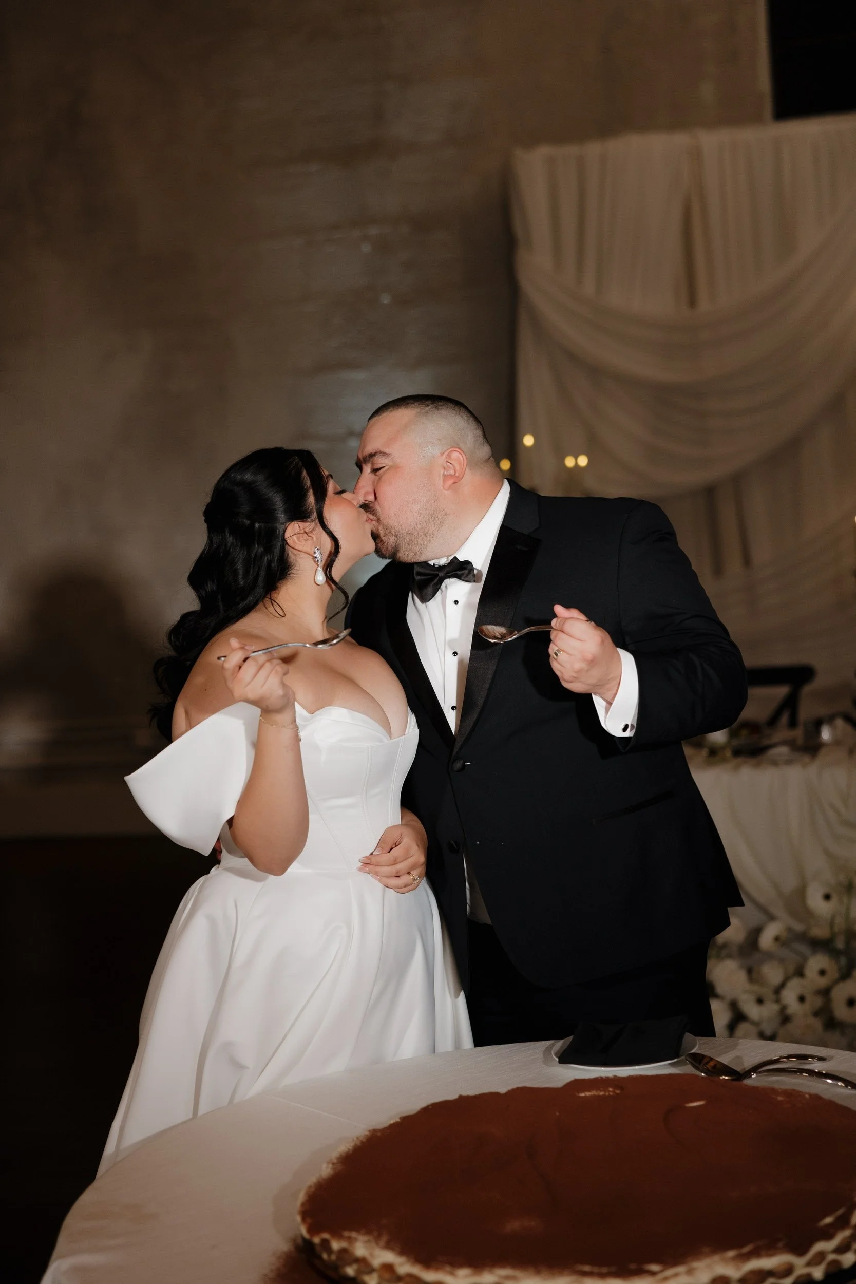 A bride and groom kiss while holding spoons at their wedding reception, with a large chocolate cake in the foreground and elegant drapery in the background.