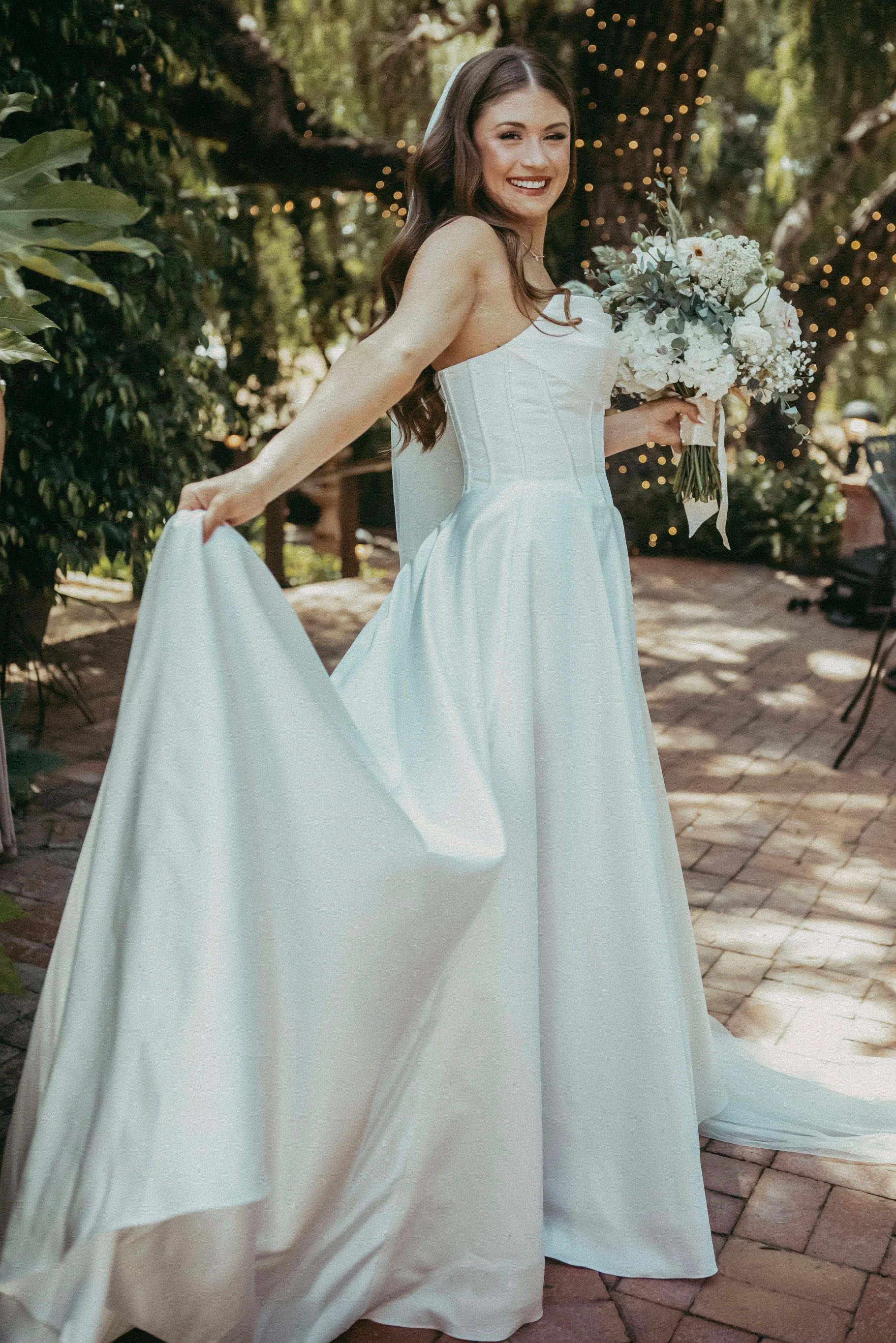 A bride in a white strapless wedding dress smiling and holding a bouquet of white flowers outdoors during the daytime.
