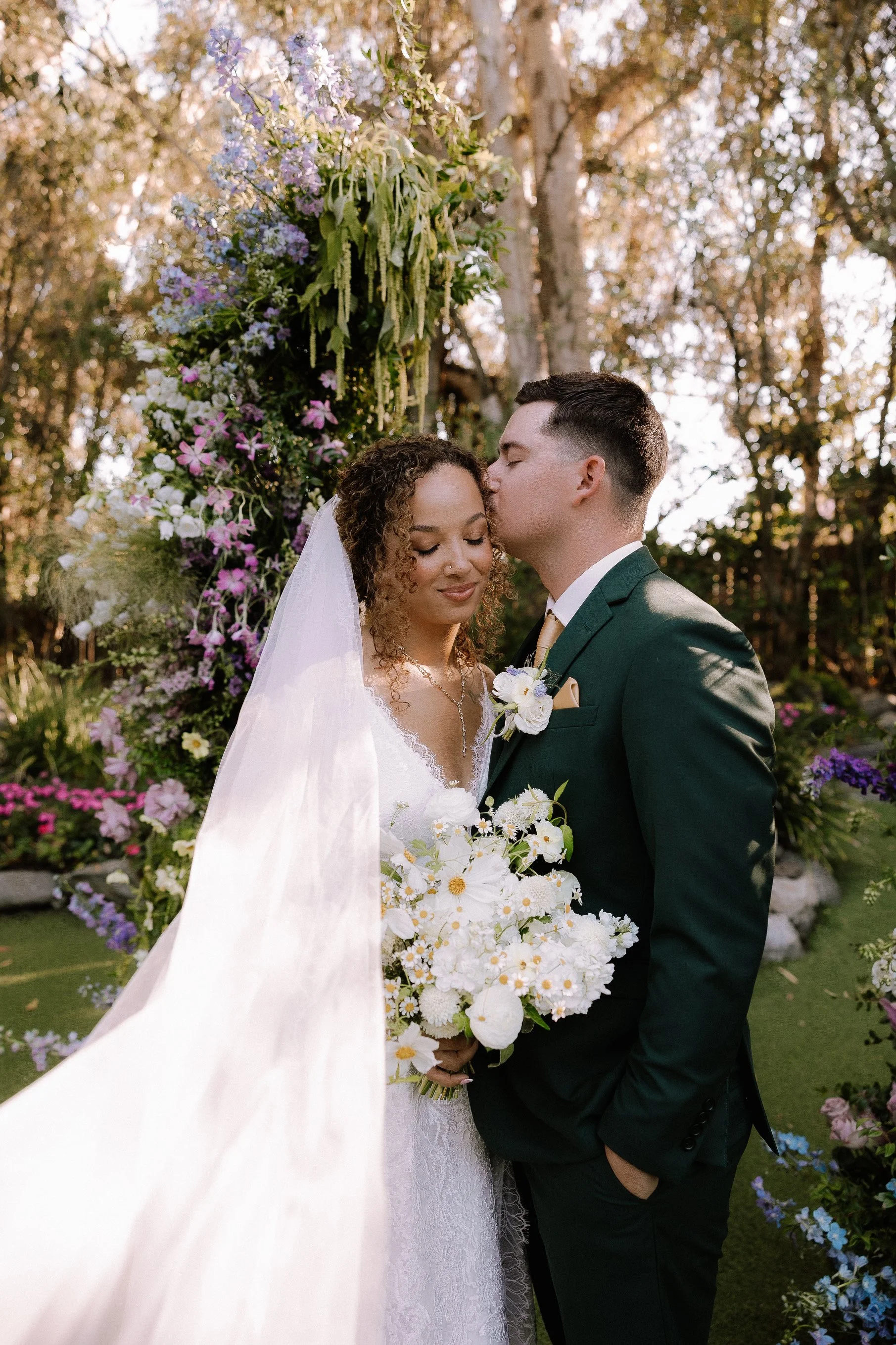 A bride and groom at their wedding, standing close together outdoors. The bride is holding a bouquet of white flowers, and the groom is kissing her forehead. They are dressed in formal wedding attire, with the bride in a white gown and veil, and the 