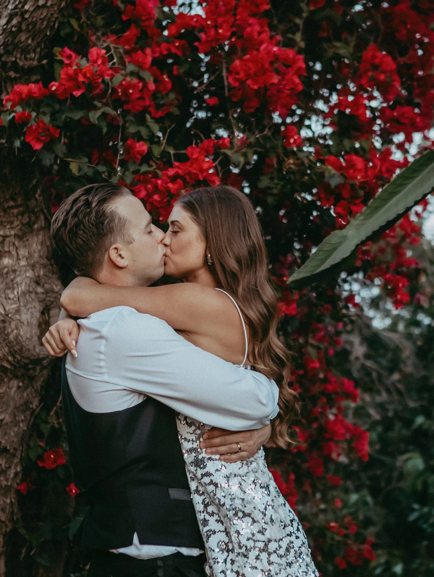A couple is embracing and kissing in front of a tree with red flowers. The woman has long, wavy brown hair and is wearing a sleeveless dress. The man has short, slicked-back hair and is dressed in a white shirt with a black vest.